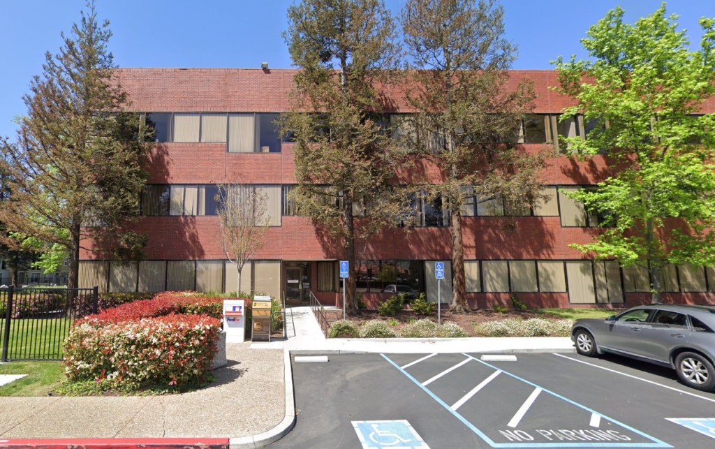 Work address of the accused, a red brick building with large windows and trees in front, with a parking lot and a gray car on the right.